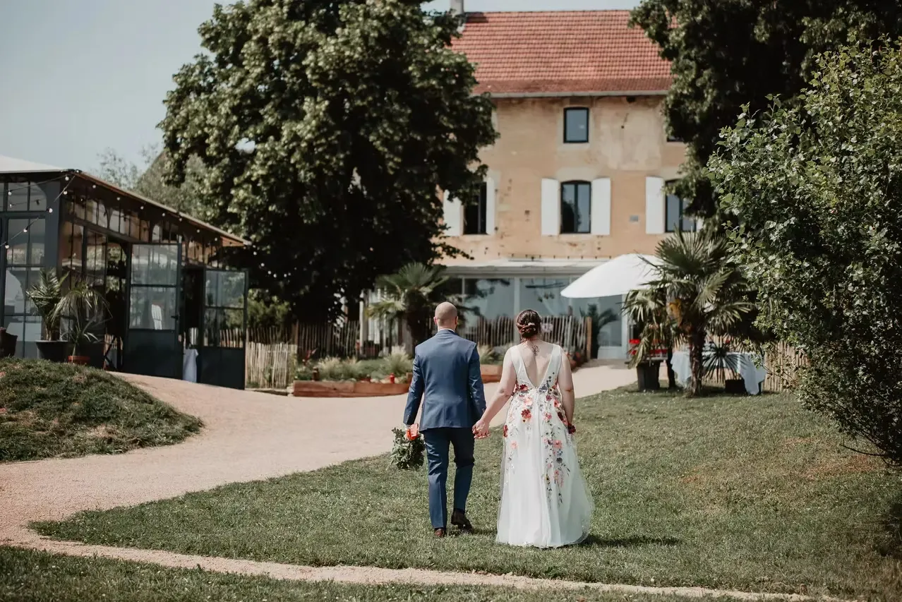 Couple de mariés marchant dans le jardin du Domaine des Plagnes en direction de l'orangerie et de la bâtisse en pierre