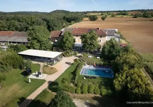 Vue aérienne du Domaine des Plagnes à Saint-Hilaire-de-Brens : bâtisse en pierre du XVIIe siècle, tente de réception et piscine entourées de champs et de verdure en Isère.