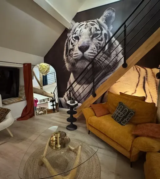 Salon du gîte Les Tilleuls avec canapé ocre, escalier en bois et fresque murale d'un tigre blanc