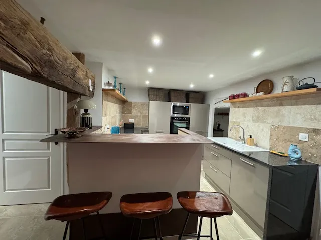 Cuisine ouverte avec îlot central, tabourets en cuir, mur en pierre et poutre ancienne