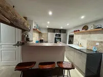 Cuisine ouverte d’un gîte du Domaine des Plagnes avec îlot central, tabourets en cuir, mur en pierre et poutre ancienne.
