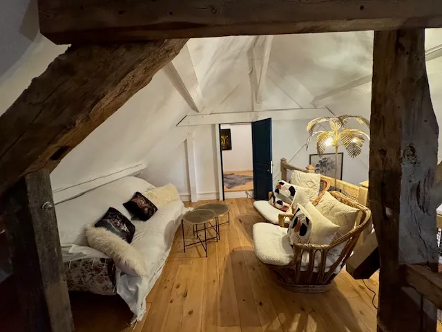 Espace détente mansardé du gîte Les Tilleuls avec poutres anciennes, banquette blanche et fauteuil en rotin