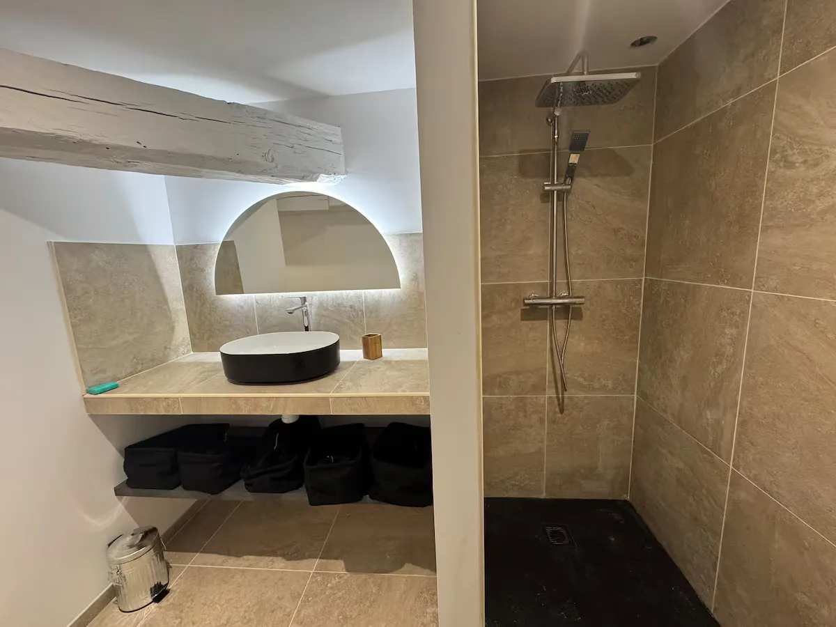 Salle d'eau mansardée avec vasque noire, miroir rond rétroéclairé et douche ouverte